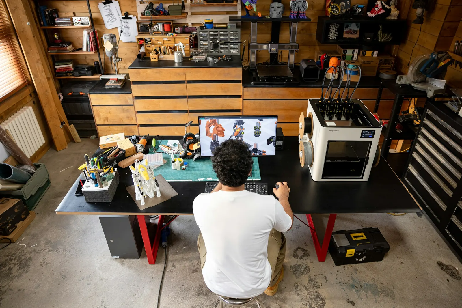 Maker designing a 3D model on a laptop beside a desktop 3D printer in a workshop