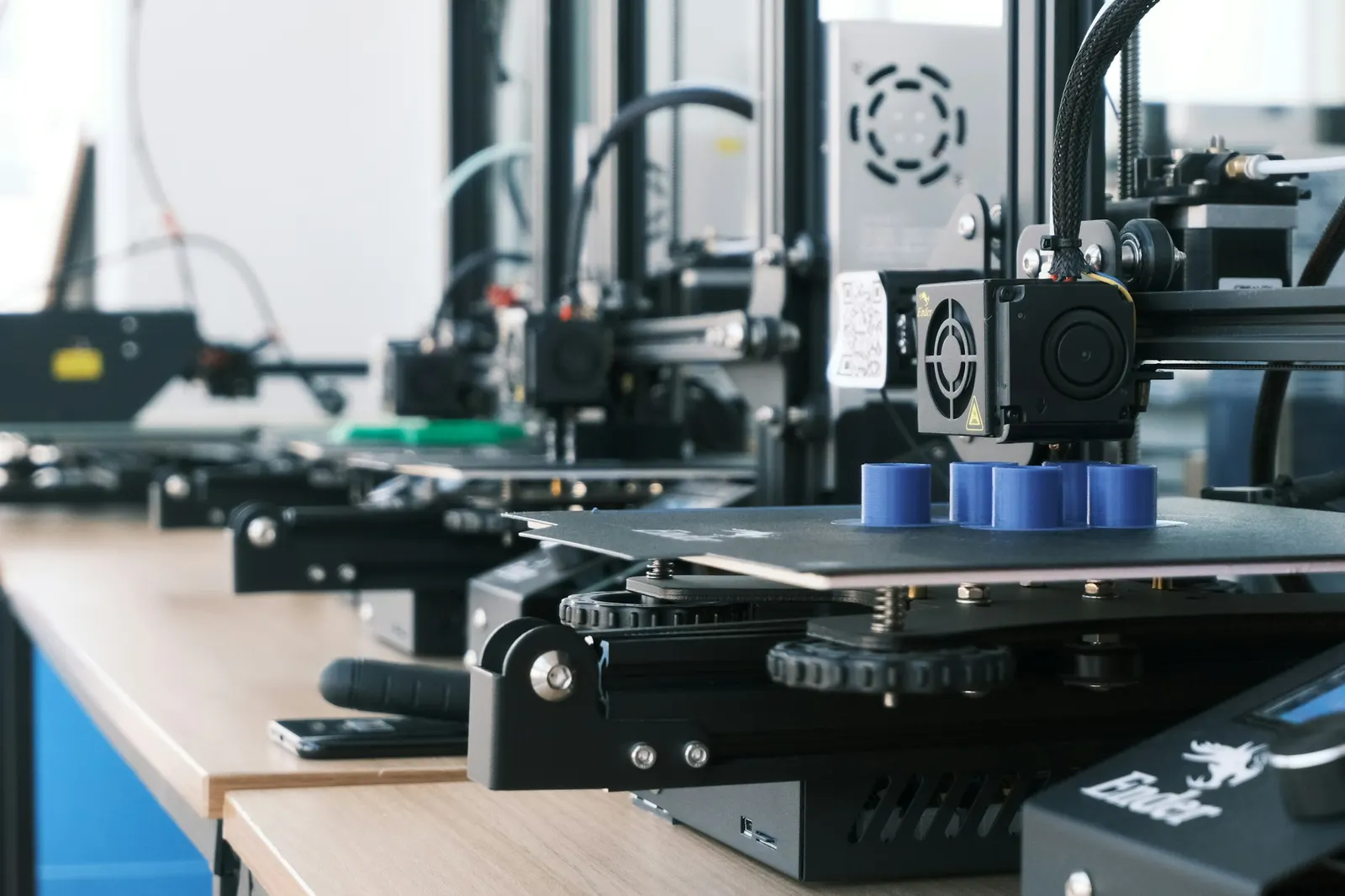 Row of 3D printers producing blue plastic parts on a workshop bench