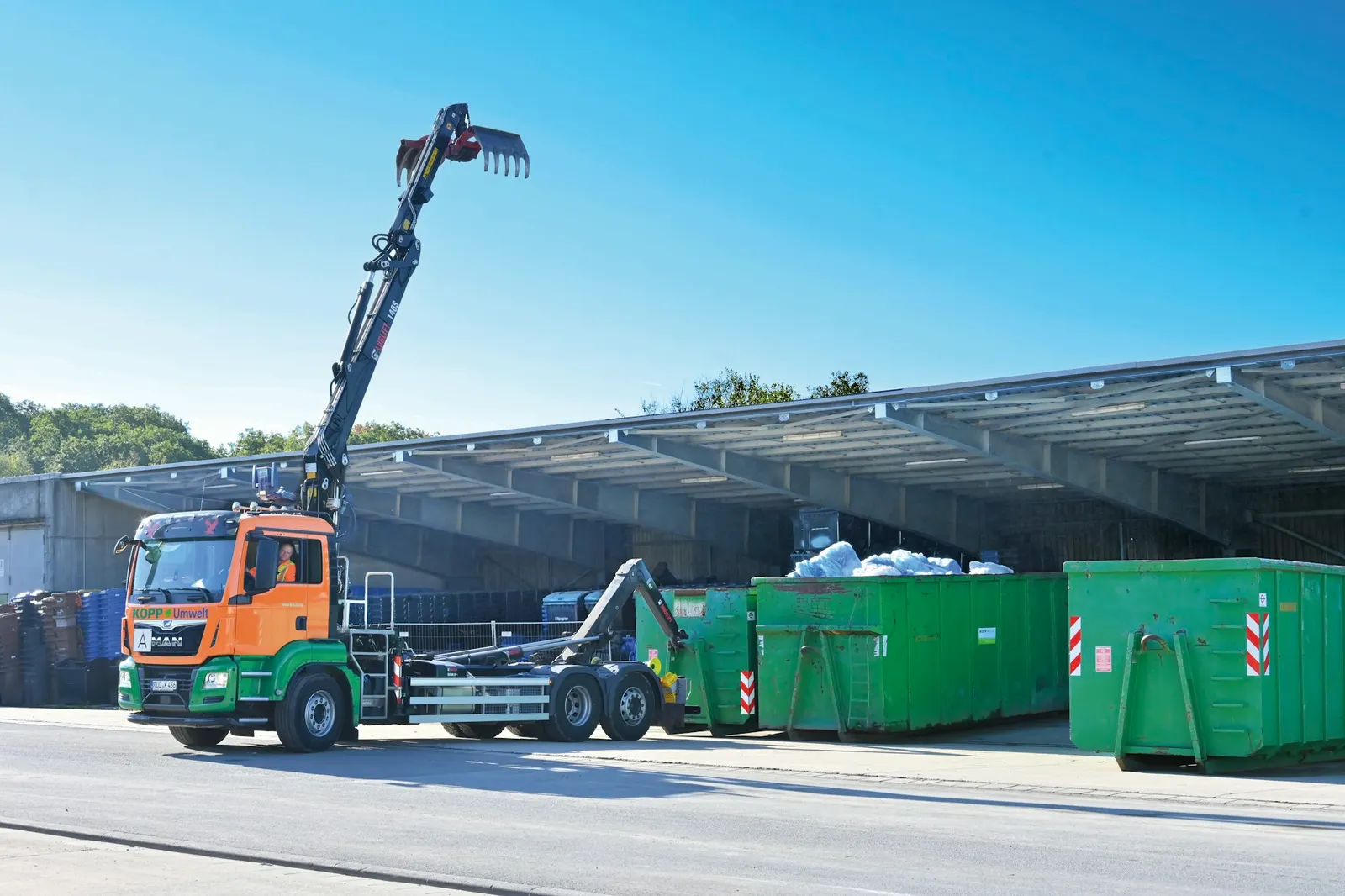 Hook-lift roll-off truck positioning a container beside green dumpsters at a waste yard
