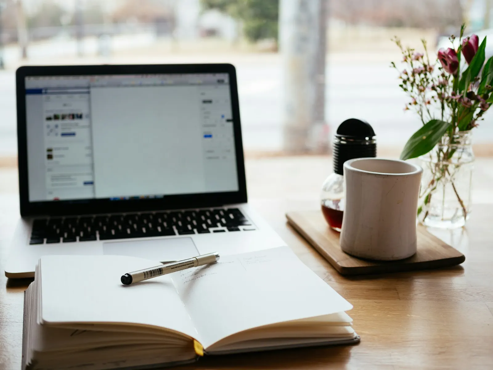 Open notebook and pen beside a laptop on a desk by a window