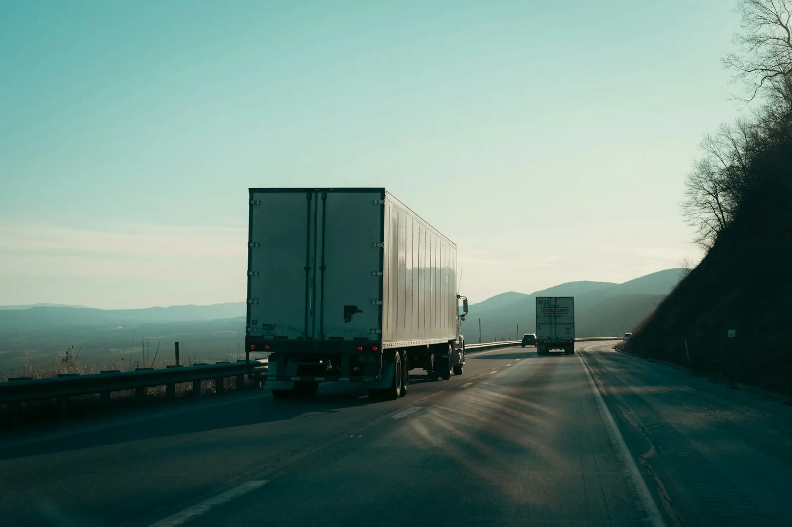 Two white semi trucks hauling trailers along a sunlit interstate highway