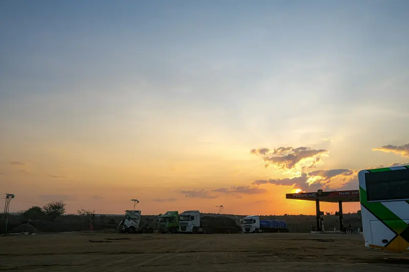 Semi trucks and a bus parked at a rest stop under a golden sunset sky