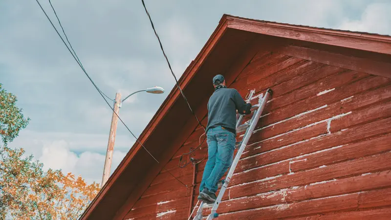 Worker on a tall ladder reaching the roofline of a red wooden house