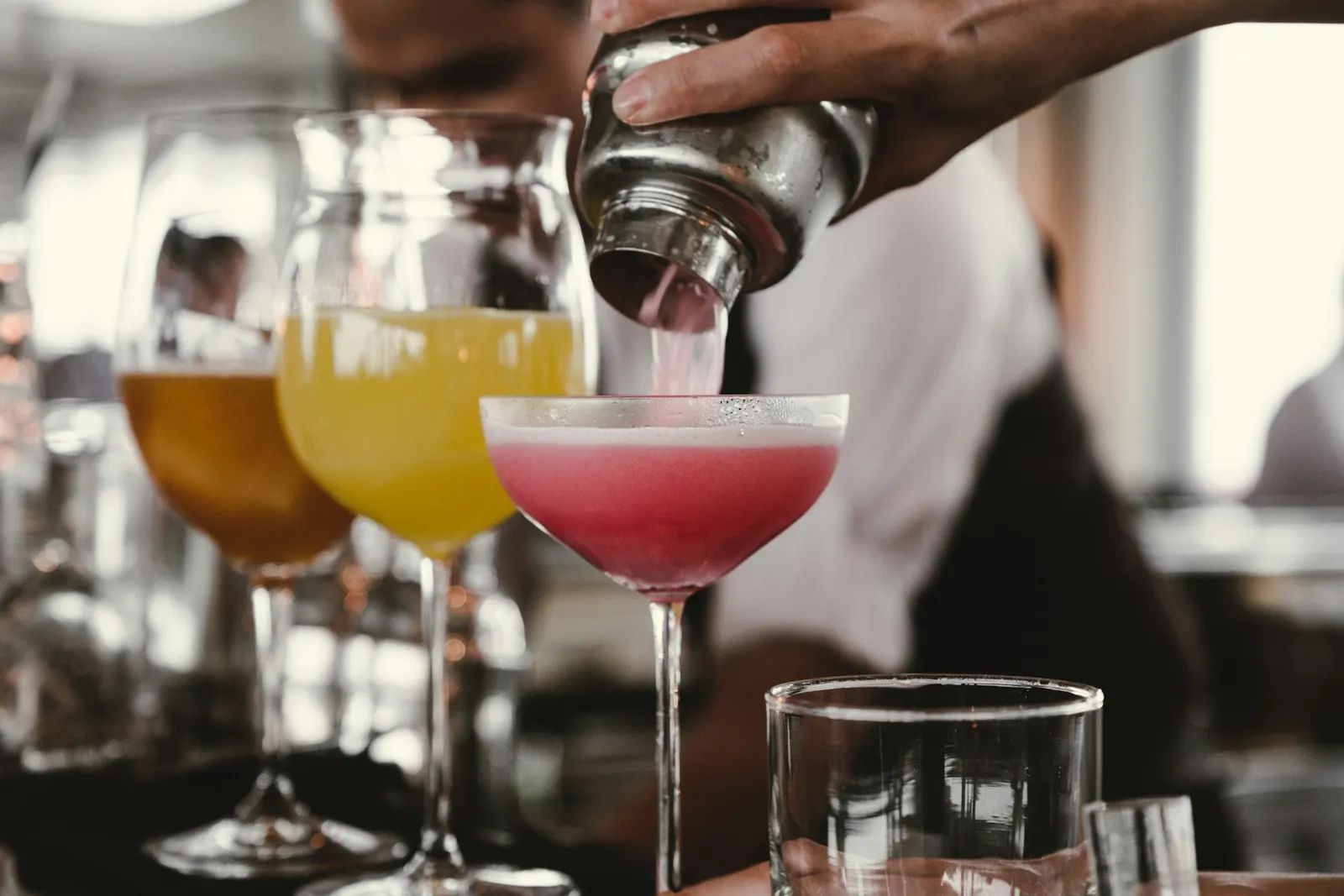 Bartender straining a pink cocktail from a metal shaker into a coupe glass