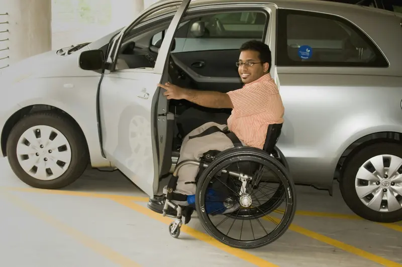 Man in a wheelchair beside the open door of a car in an accessible parking bay