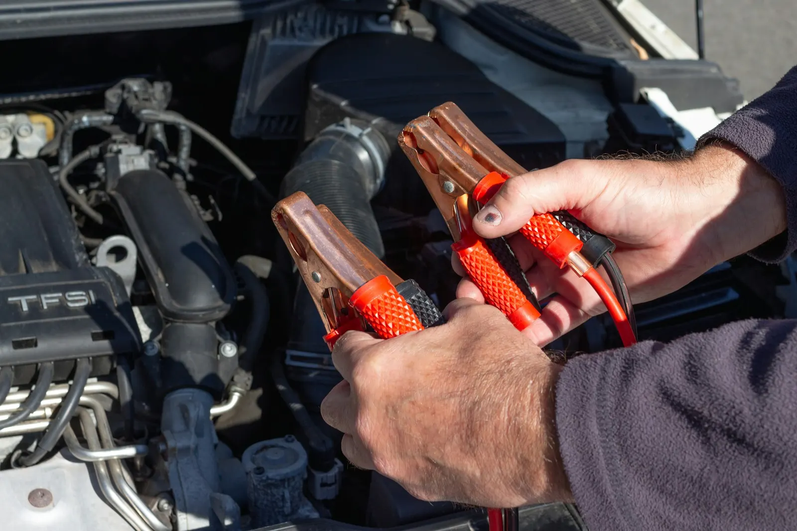 Hands holding red and black jumper cable clamps over an open car engine