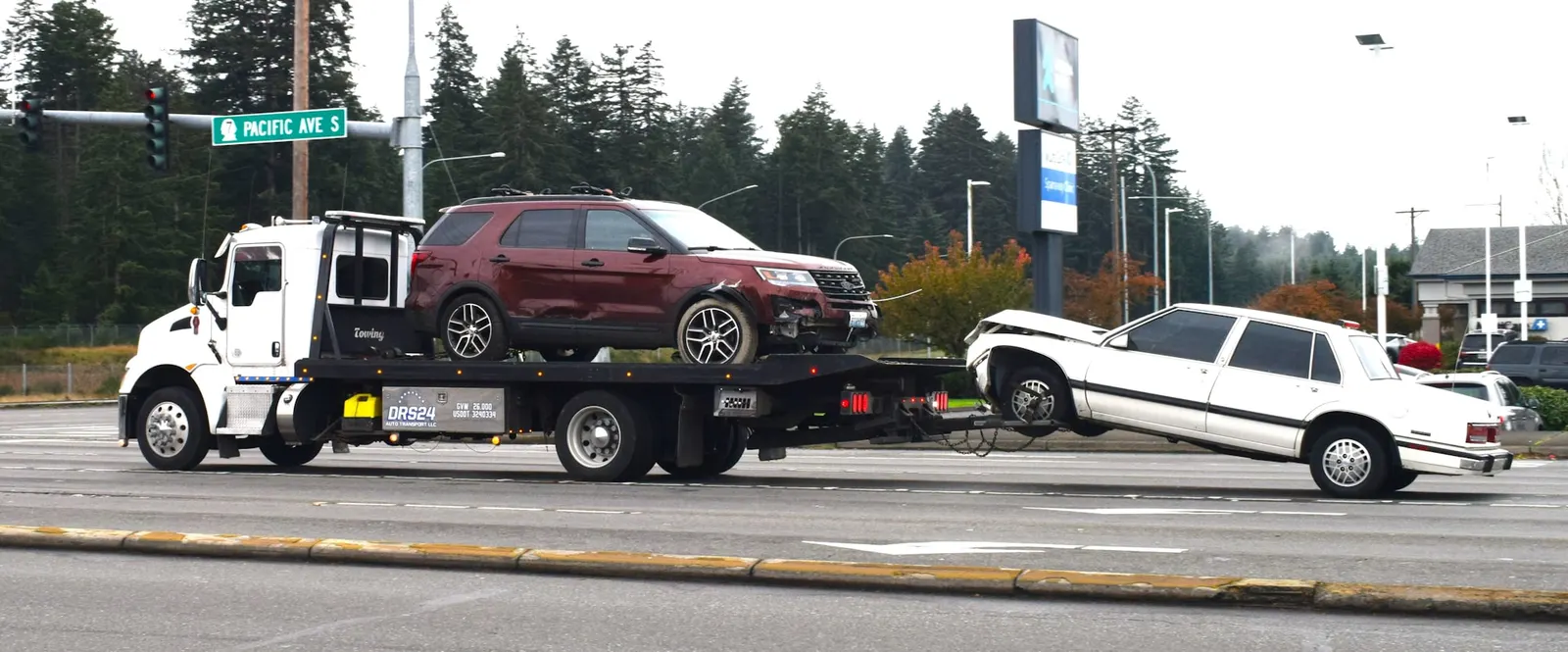 Flatbed service truck hauling an SUV and towing a second car along a city road