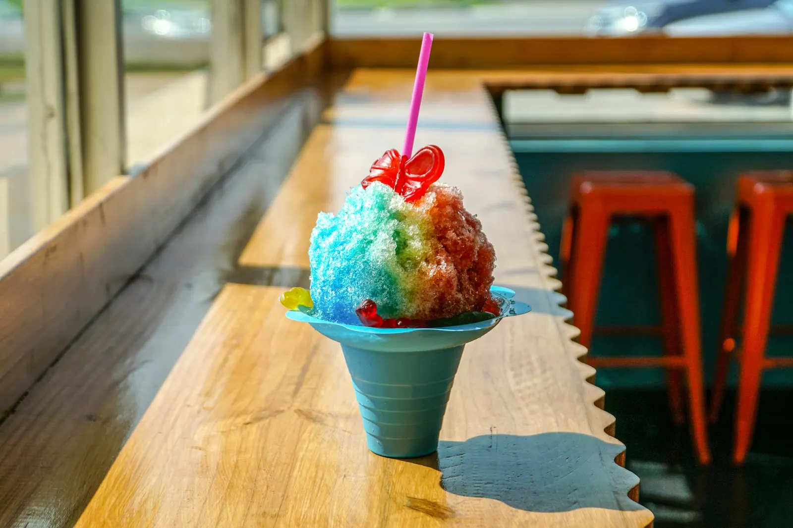 Rainbow shaved ice piled high in a blue flower cup with a straw and gummy topping