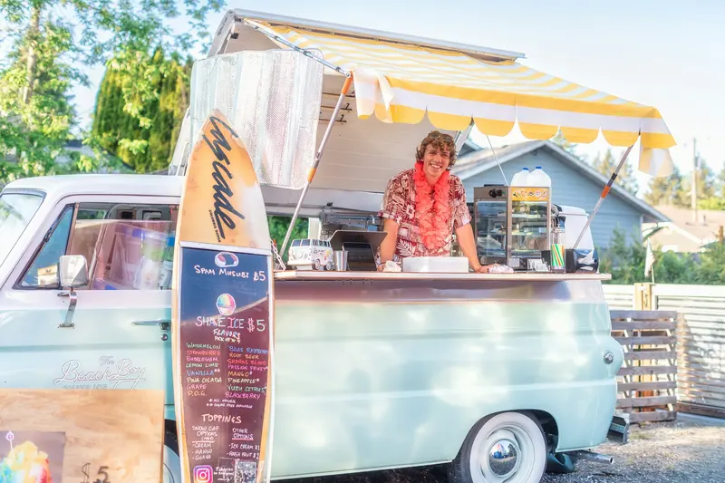 Smiling vendor serving shave ice from a vintage van with a striped awning on a sunny day