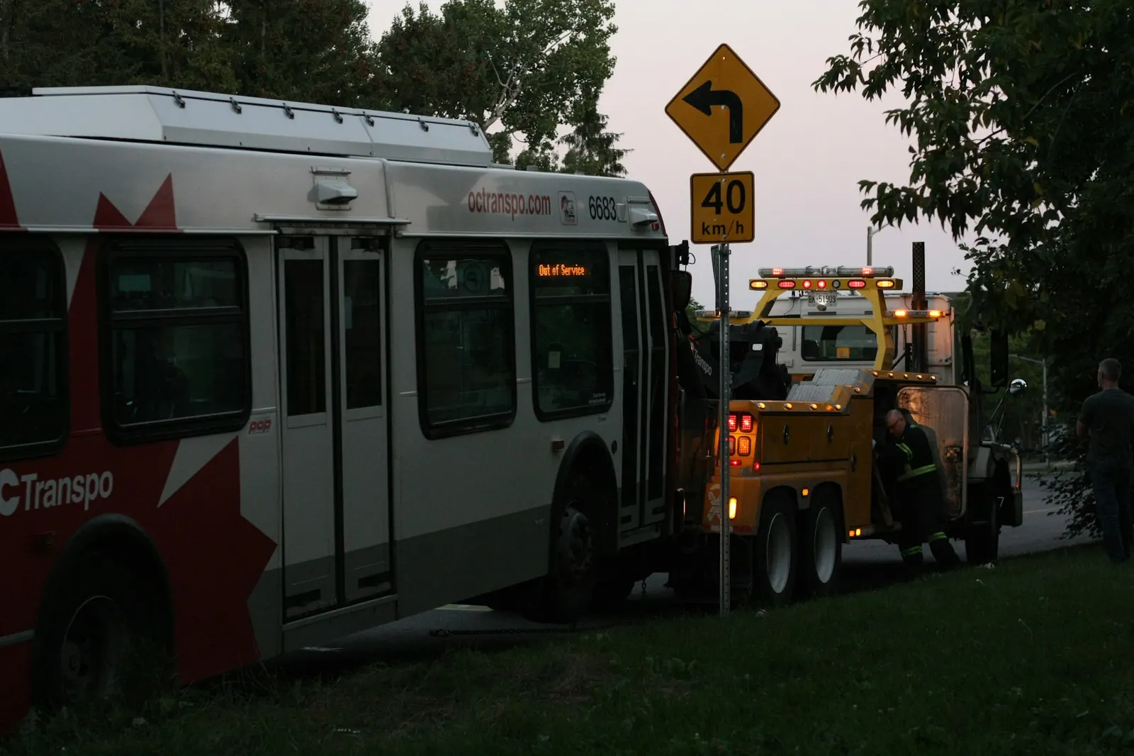 Yellow wrecker with amber lights towing a bus at dusk beside the road