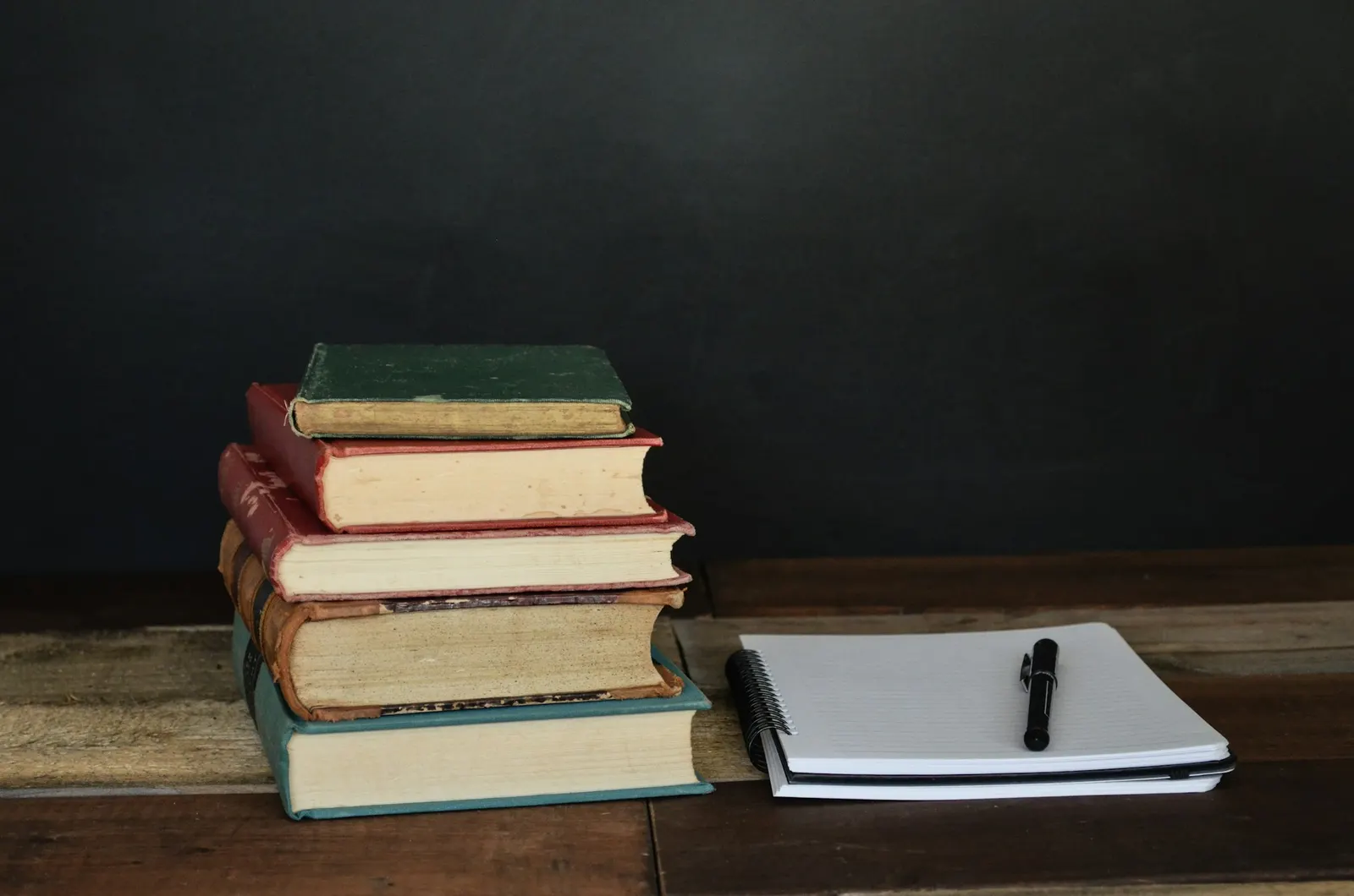 Stack of hardcover textbooks beside a notebook and pen on a wooden desk