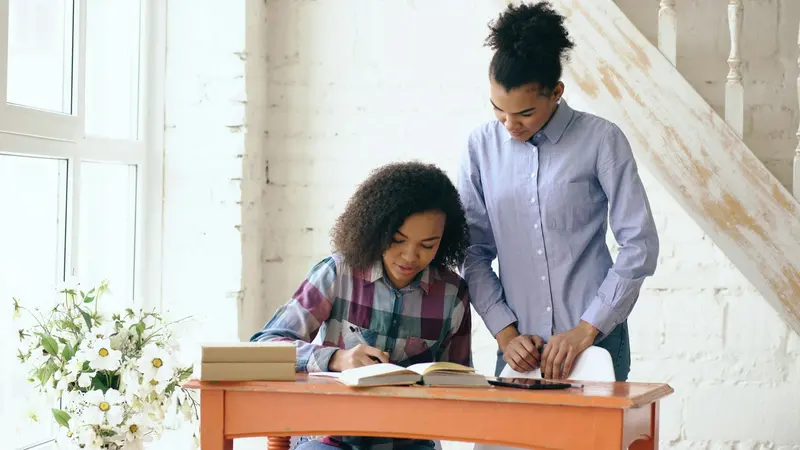 Tutor leaning over a teenage student working through homework at a sunlit table