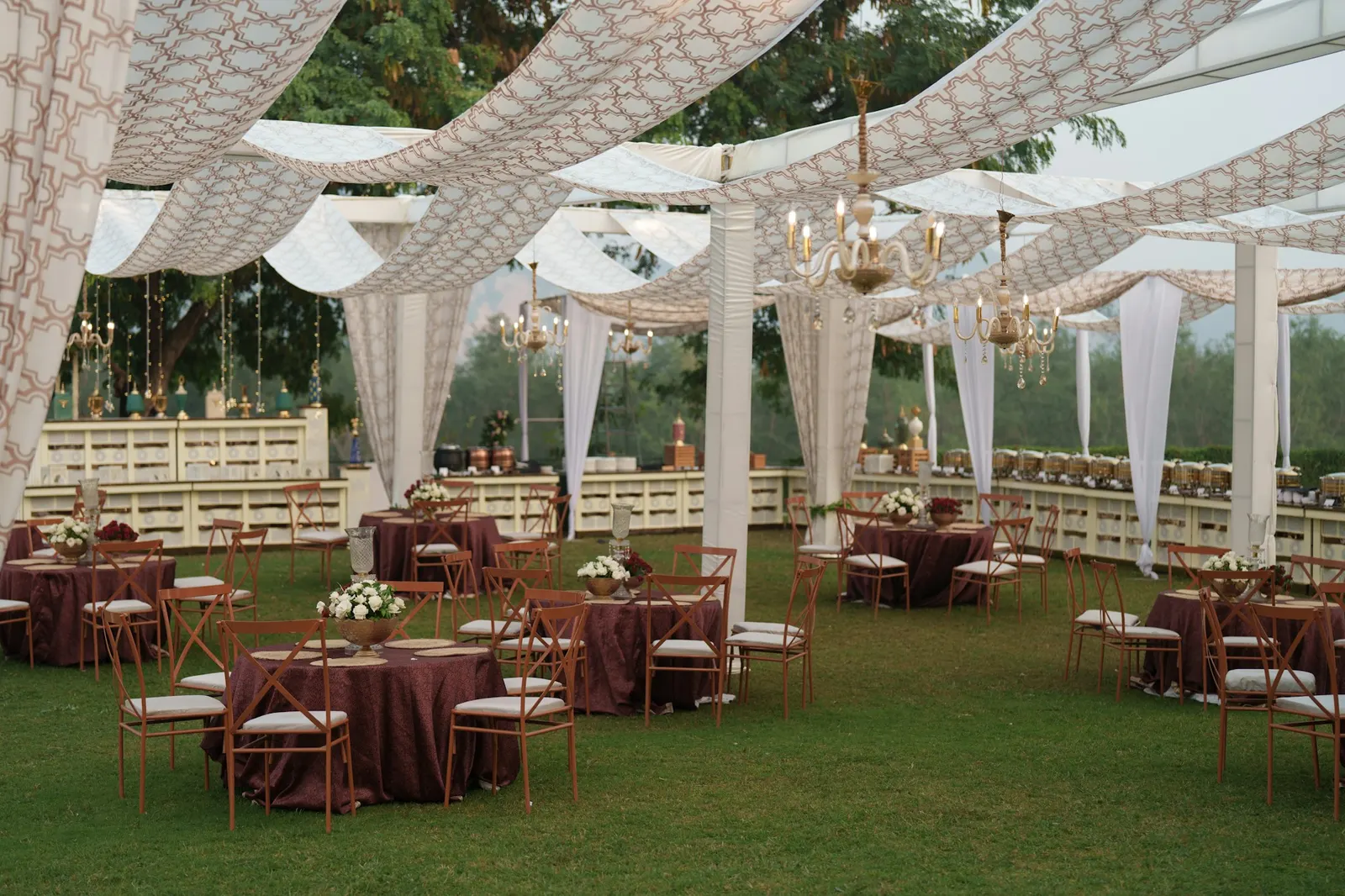 Elegant outdoor reception setup with draped canopy, chandeliers and round tables